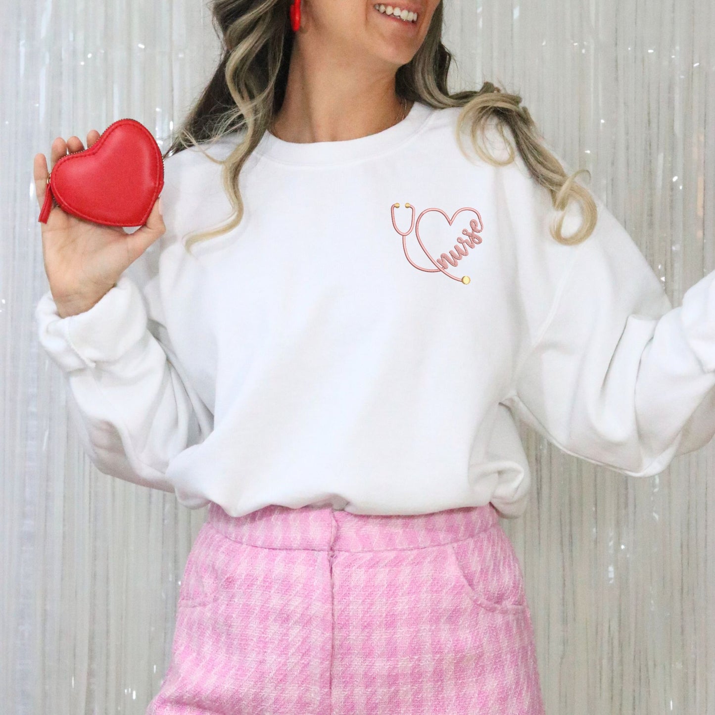 Woman wearing a white sweatshirt with a stethoscope heart design, holding a red heart-shaped purse against a light background.