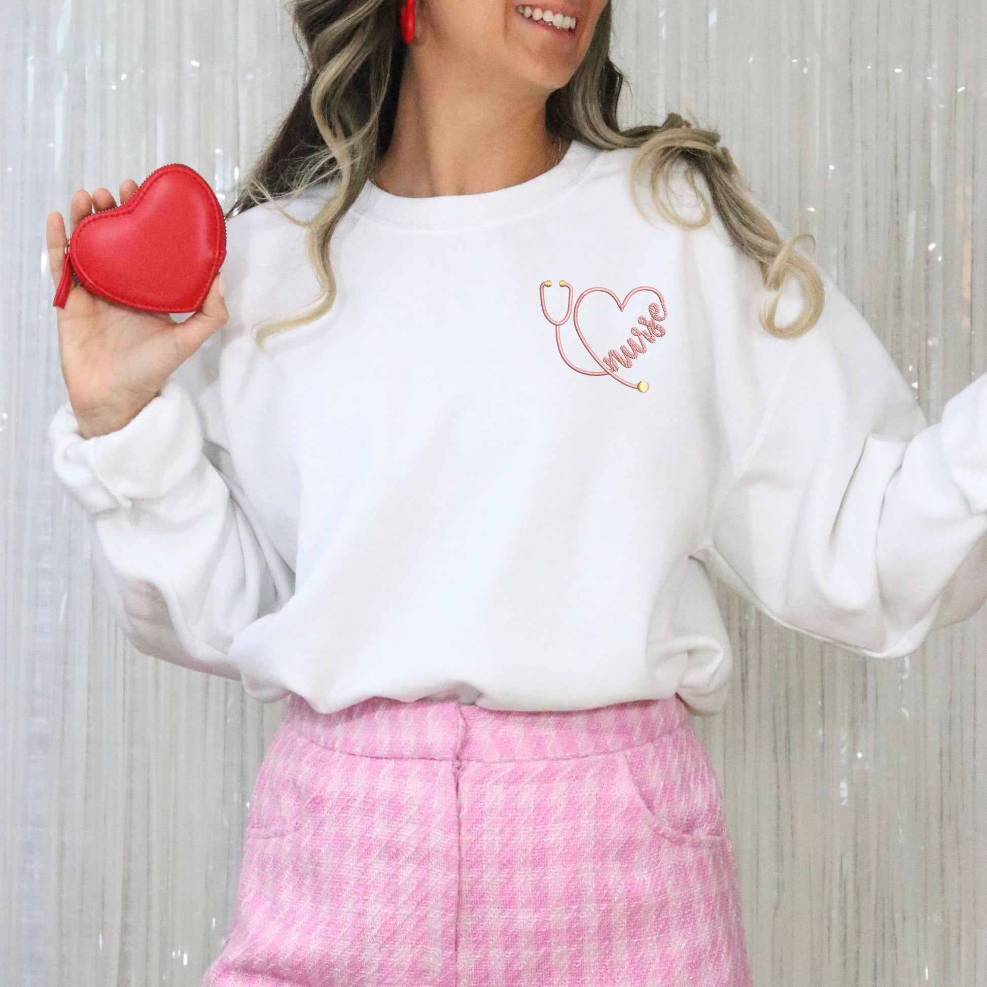 Woman wearing a white sweatshirt with a stethoscope heart design, holding a red heart-shaped purse against a light background.
