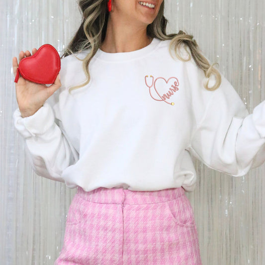 Woman wearing a white sweatshirt with a stethoscope heart design, holding a red heart-shaped purse against a light background.