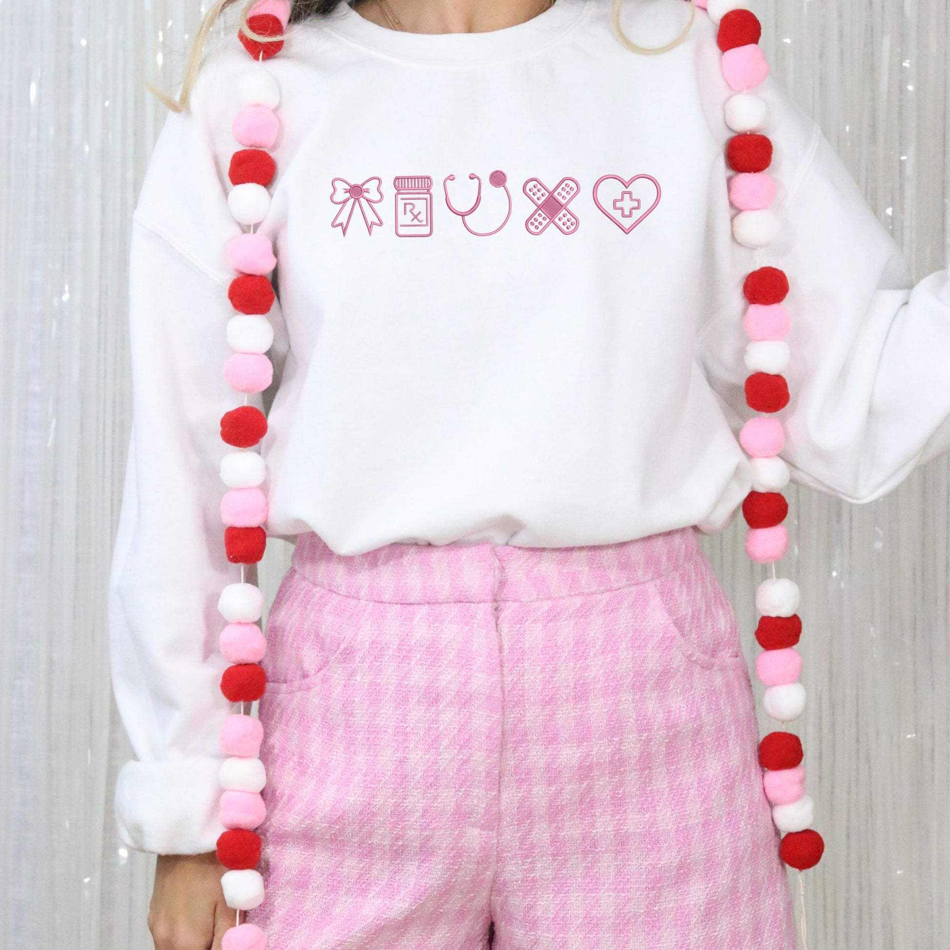 Person wearing a white sweatshirt with pink text and pink pants, holding a long decorative garland. Design is a bow, Rx bottle, stethoscope, bandaids & a heart embroidered across the chest