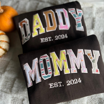 Two black shirts with 'DADDY EST. 2024' and 'Mommy EST. 2024' text on a textured surface with pumpkins.